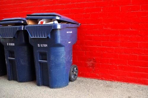 Two large dark blue wheelie bins made of high-density plastic with textured surfaces and fitted with black lids, positioned on a concrete pavement beside a vibrant red brick wall. The bins are placed close together, with one slightly leaning against the other, and contain visible rubbish, including a cardboard box and various waste items protruding from the open lid of one bin. The scene appears to be outside a commercial or industrial building, with the bins used for rubbish collection or waste removal services in Southgate. The background features the brick wall's uniform pattern and the paved surface is clean with some minor gravel and dirt visible around the bin wheels, indicating typical outdoor storage of waste containers.