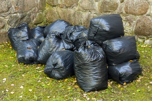 A collection of approximately ten black plastic rubbish bags, filled and tightly knotted at the top, are arranged in a loose cluster on a grassy outdoor area. The bags are situated in front of a roughly textured stone wall composed of irregularly shaped stones with various shades of brown, grey, and beige, some moss or lichen growth visible. The grass beneath the bags shows patches of green with small scattered leaves and debris. The lighting appears natural, suggesting an outdoor setting during daytime. This scene depicts typical waste collection for rubbish removal services, potentially associated with residential or small-scale commercial waste disposal in Southgate or nearby areas, as part of a local rubbish clearance effort handled by Business Waste Removal Southgate.