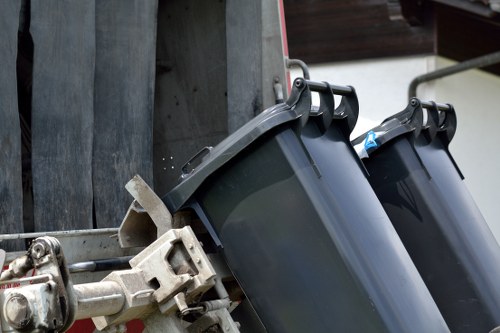 A close-up view of two black plastic wheeled trash bins attached to a metal waste collection cart, positioned on a driveway or pavement surface. The bins are made of durable, smooth plastic with slightly textured surfaces, and are aligned side by side with their lids closed. One bin has a blue handle for lifting, and both are secured to a metallic framework that attaches to the cart, which appears to be used for waste collection or rubbish removal services. The background features a wooden fence or wall, indicating an outdoor setting typical of a residential or commercial rubbish collection point. The lighting suggests natural daylight, highlighting the clean appearance of the bins and their functional design. This scene depicts equipment used by waste removal services in Southgate, supporting local rubbish disposal needs.