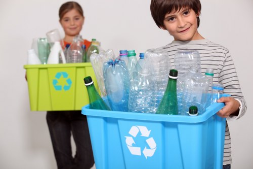 A young boy with short dark hair, wearing a grey and white striped long-sleeve shirt, is holding a light blue recycling bin filled with empty plastic bottles and containers. The bottles vary in size and shape, with some having green or clear plastic, and are arranged inside the bin, which features a white recycling symbol on the front. In the background, a young girl with shoulder-length light brown hair, dressed in a grey top, is holding a bright green recycling container filled with similar plastic bottles, standing in front of a plain white wall. The scene takes place in an indoor setting, possibly a home or a community centre, emphasizing waste separation and recycling efforts. The lighting is bright and even, ensuring clear visibility of the objects and the facial expressions of both children. This image aligns with proper rubbish management themes, illustrating responsible disposal of plastic waste as part of local waste removal initiatives, such as those provided by Business Waste Removal Southgate.