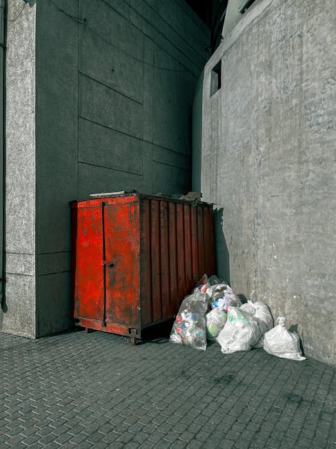 A person wearing a green long-sleeved shirt, a plaid yellow and grey jacket, and grey trousers is holding open a large black rubbish bag lined with a silver interior, positioned outdoors on a grassy a