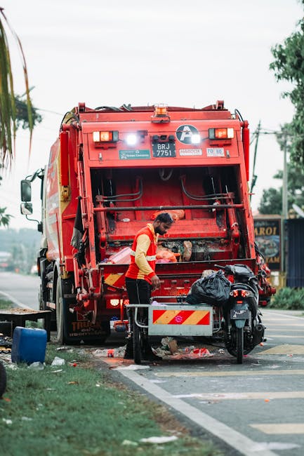 A waste collection worker, dressed in a yellow high-visibility vest and black trousers, is positioned at the rear of a large red rubbish truck, which is parked on the side of a street. The worker is a