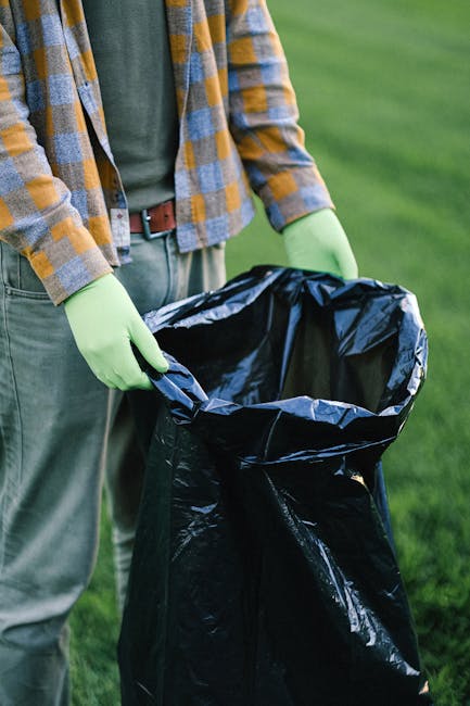 A waste collection worker, dressed in a yellow high-visibility vest and black trousers, is positioned at the rear of a large red rubbish truck, which is parked on the side of a street. The worker is a